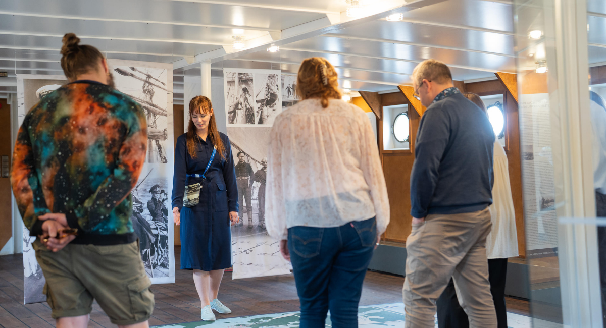 Valokuva opastuksesta, jossa opas kertoo kolmelle kuulijalle Suomen Joutsenesta.